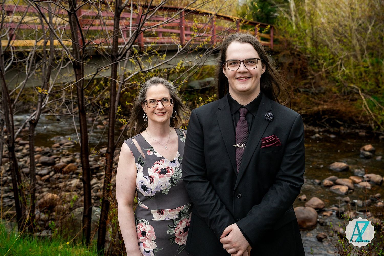 A couple wearing formal attire stands in front of a small stream and bridge during an outdoor photo session.