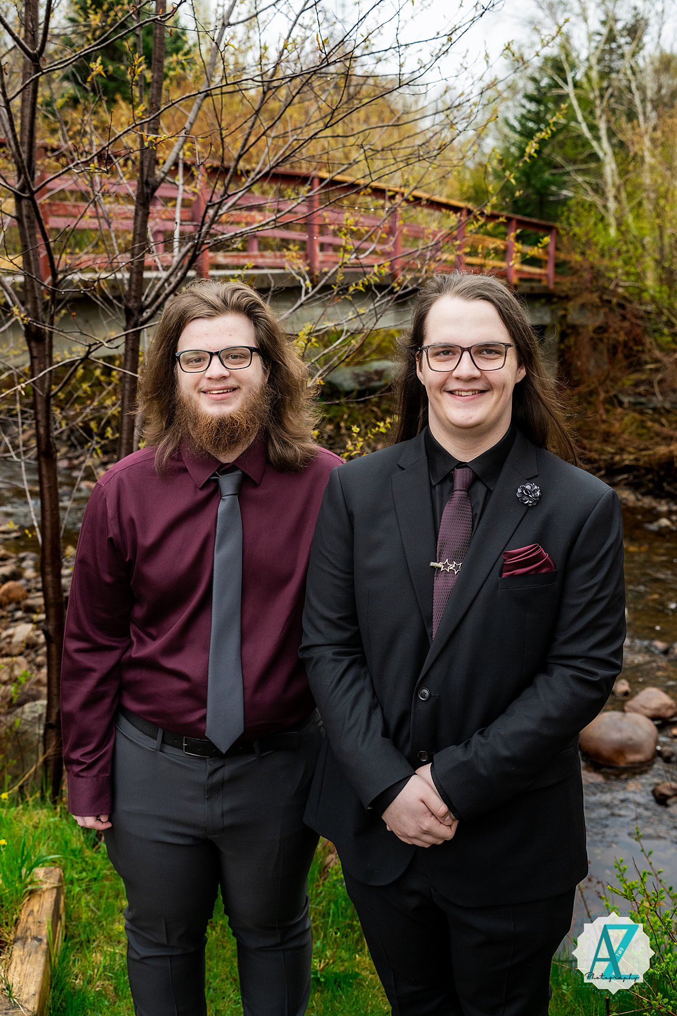 Two stylishly dressed figures stand together in front of a wooden bridge at an outdoor venue with autumn trees.