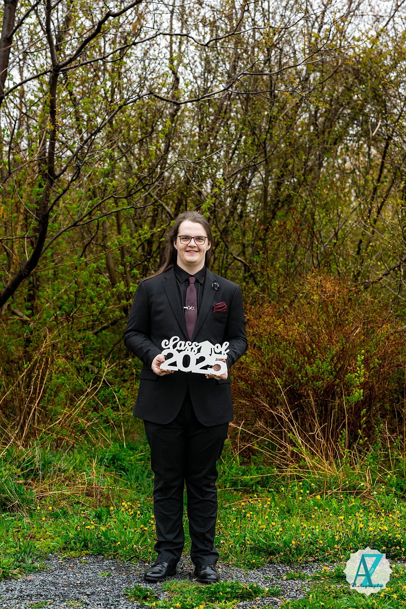 A figure in a black suit stands holding a sign against a backdrop of green foliage on a gravel path.