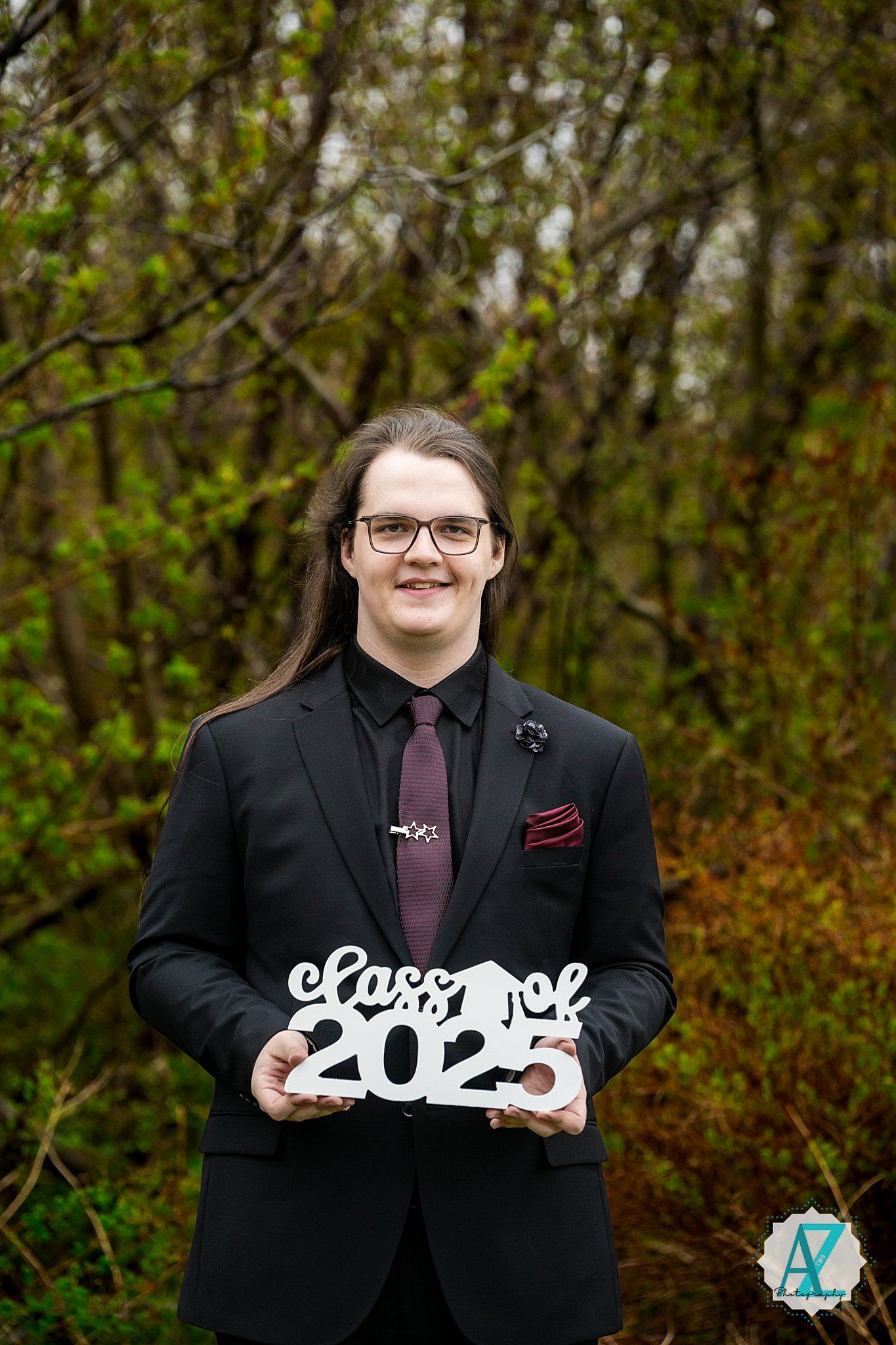 A graduate in a black suit and burgundy tie holds a decorative 'Class of 2023' sign in a natural outdoor setting.