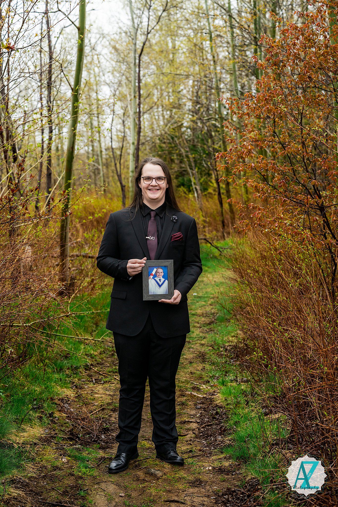 Person wearing a dark suit stands on a forest path surrounded by autumn foliage and bare trees.