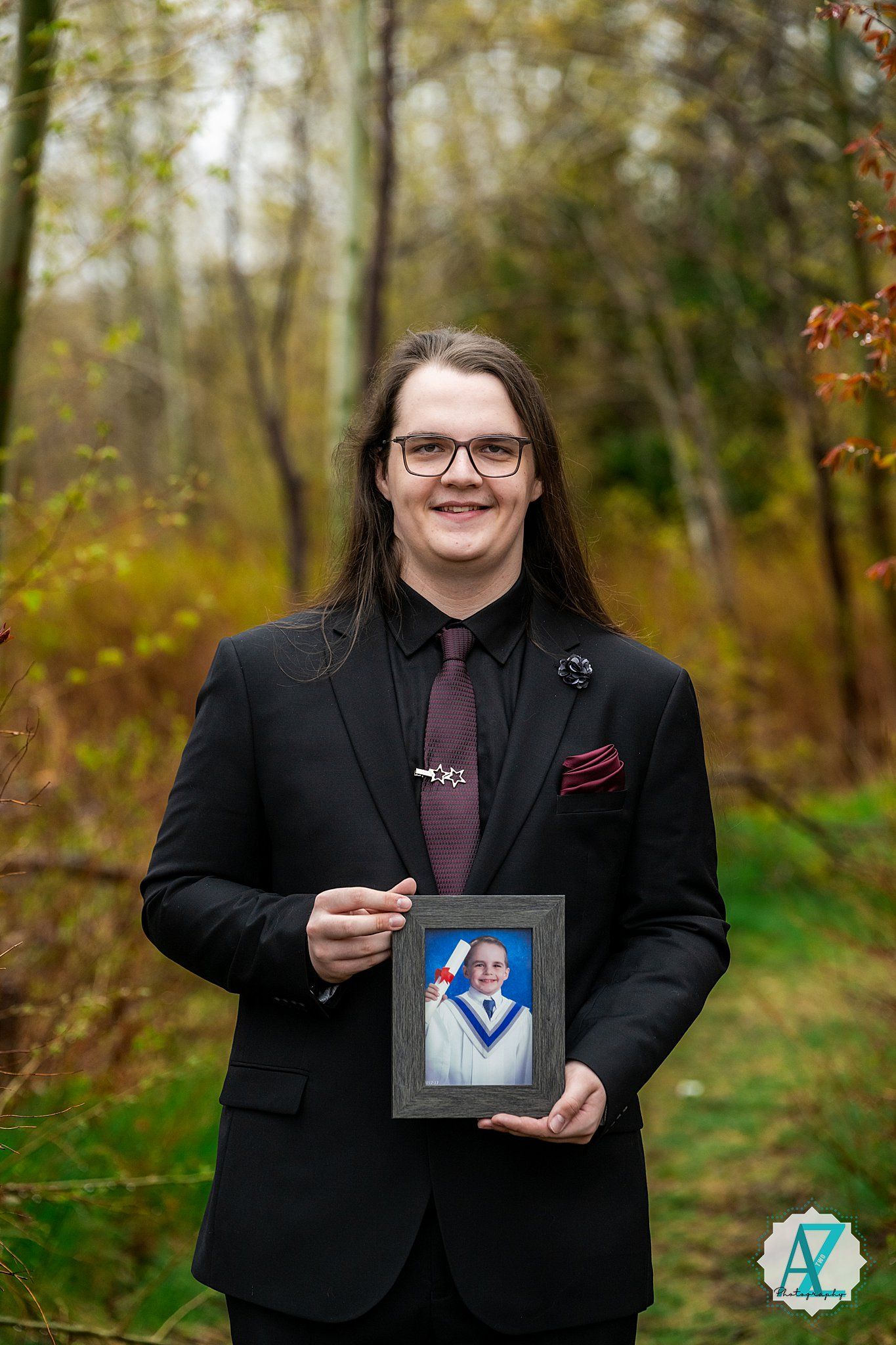 Someone in a black suit and burgundy tie holds a framed photograph while standing in an autumn forest setting.