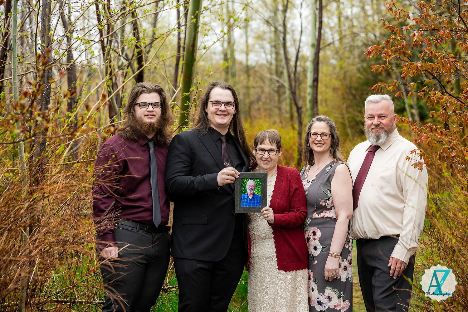 A group of people stand together in a wooded area with autumn foliage, holding a blue book.