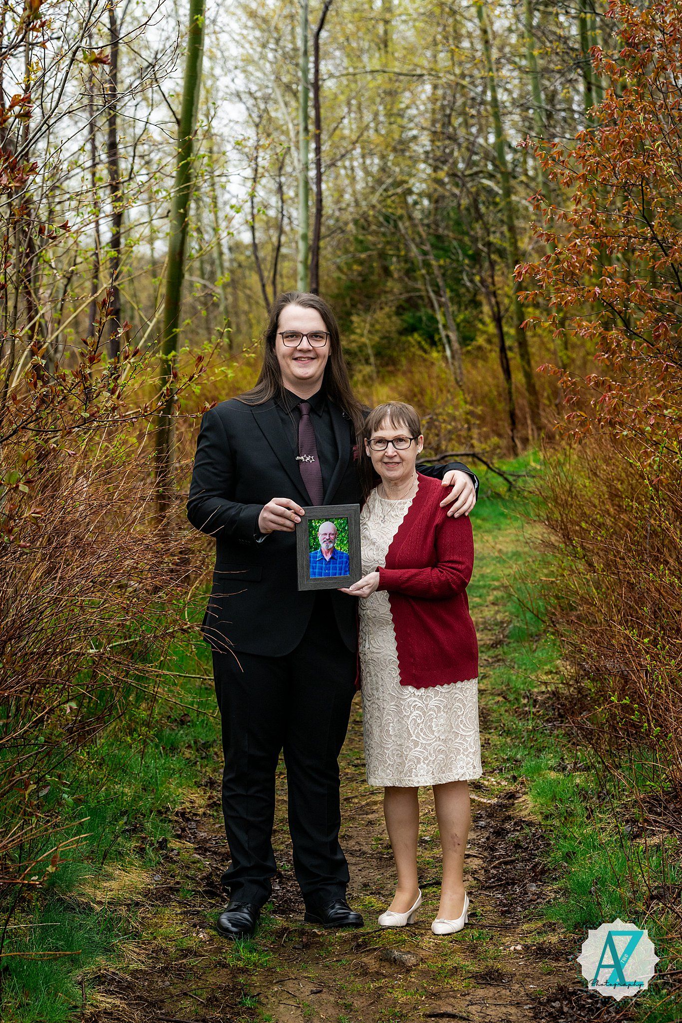 Two people stand on a forest path holding a framed photo during autumn with colorful foliage in the background.