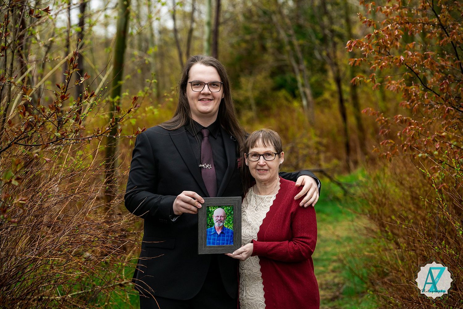 Two people stand in an autumn forest setting holding a framed photograph against a background of fall foliage.