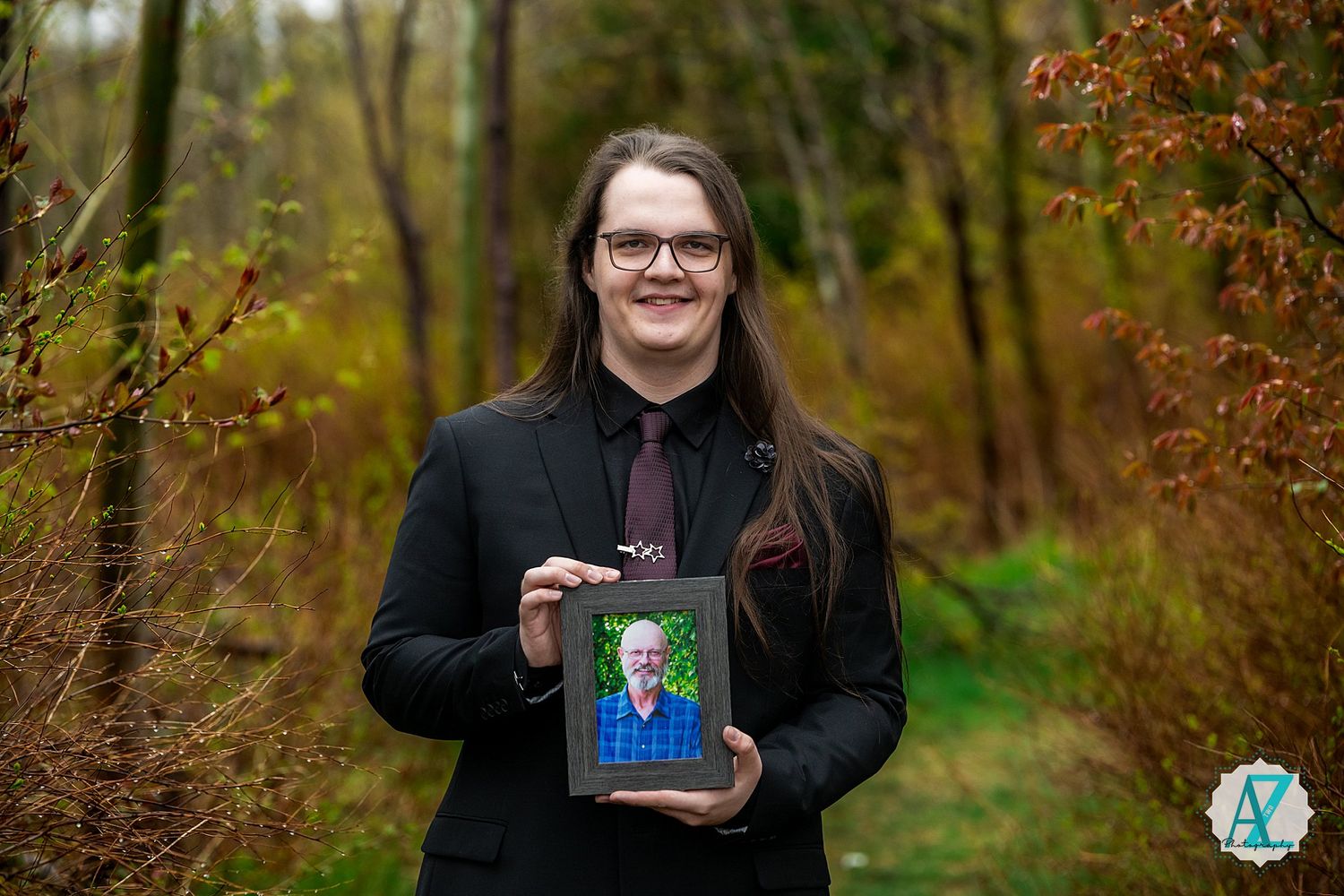 Person in black jacket stands in autumn forest holding a framed portrait photograph.
