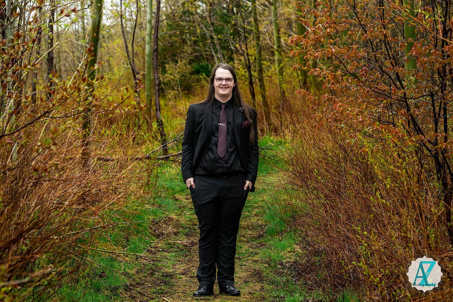 A person stands smiling on a forest trail surrounded by autumn foliage and green undergrowth.