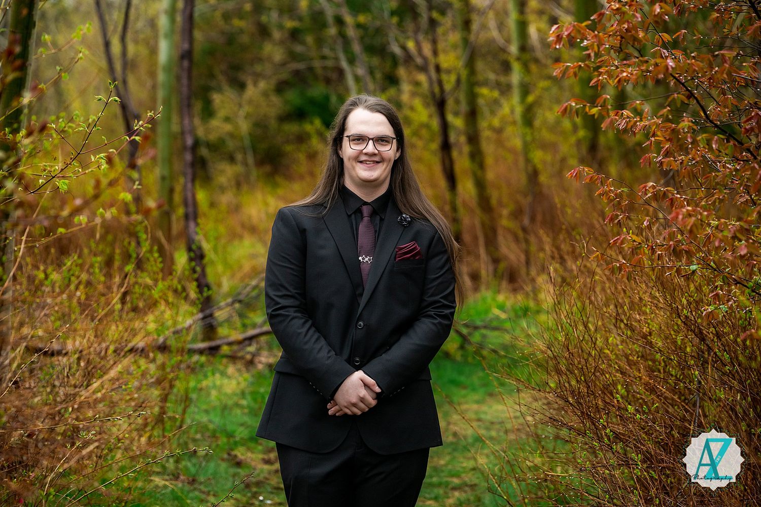 A person in a black suit and glasses stands smiling outdoors in an autumn forest setting.