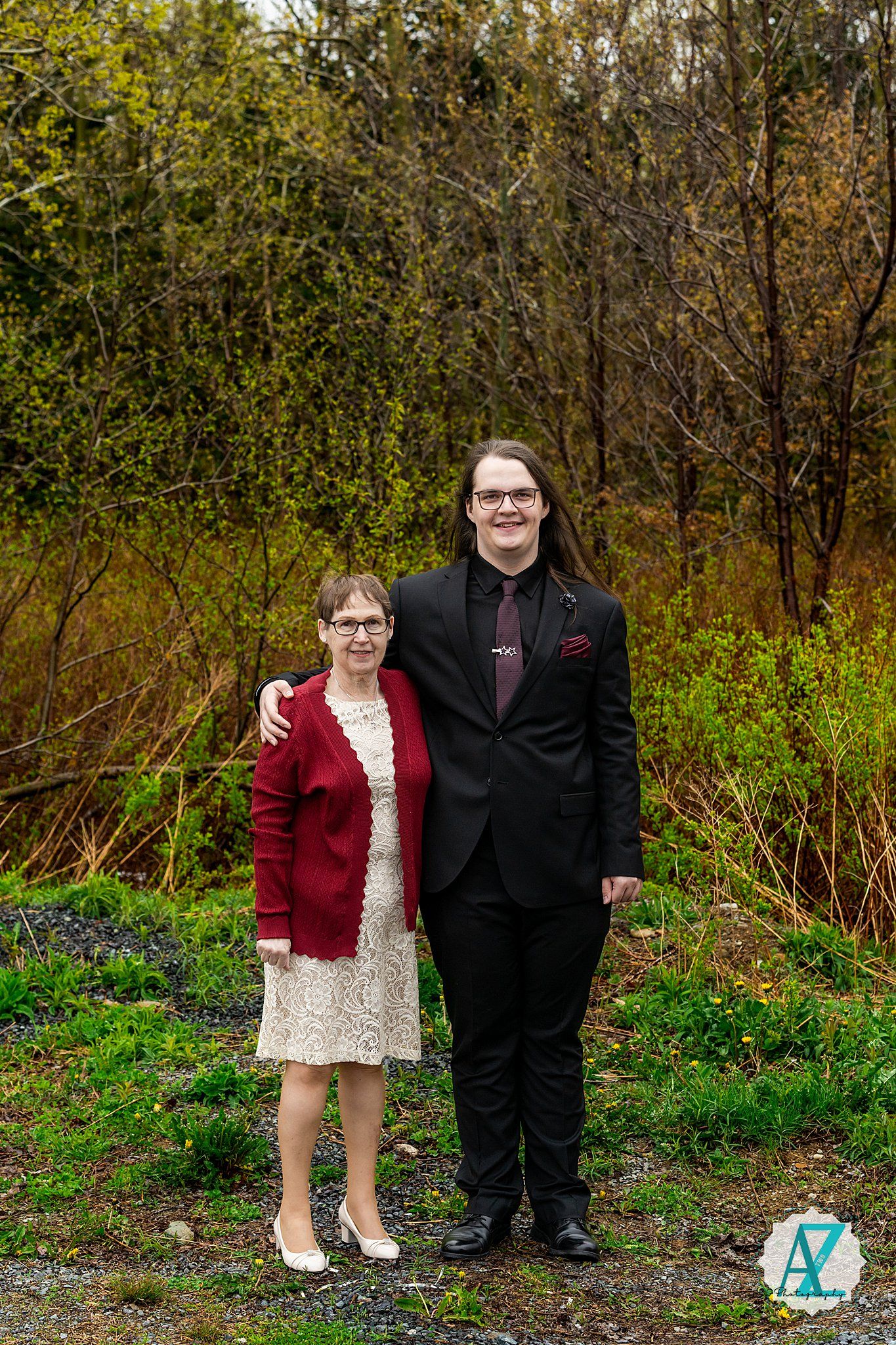 Two figures stand outdoors in a scenic natural setting with trees and greenery in the background.
