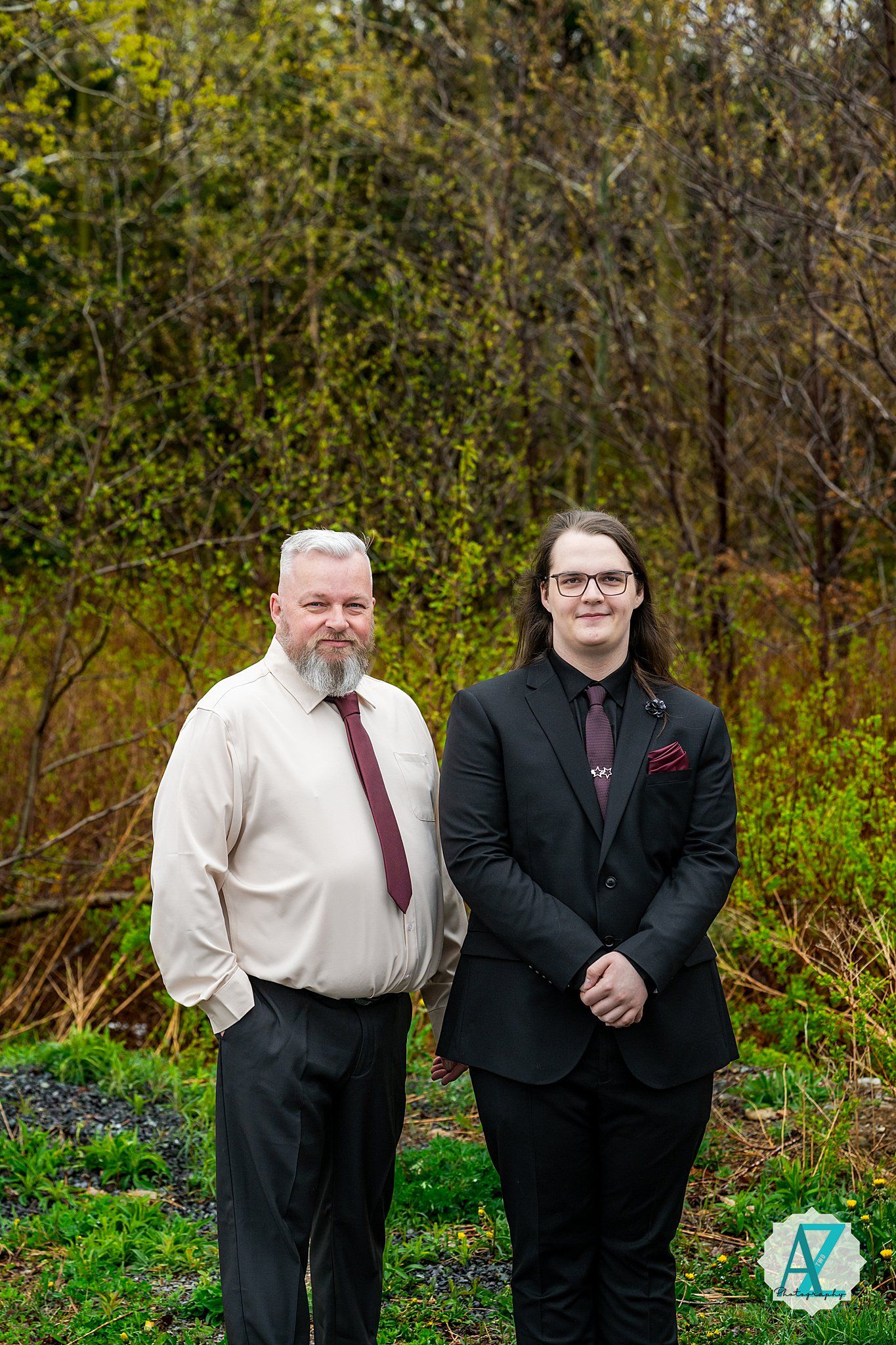 Two people in formal attire stand together outdoors in front of green foliage on a sunny day.