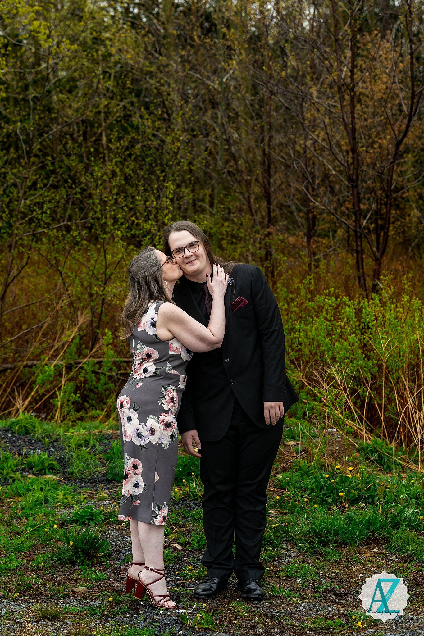 Two people in formal attire stand together outdoors with natural greenery and trees in the background.