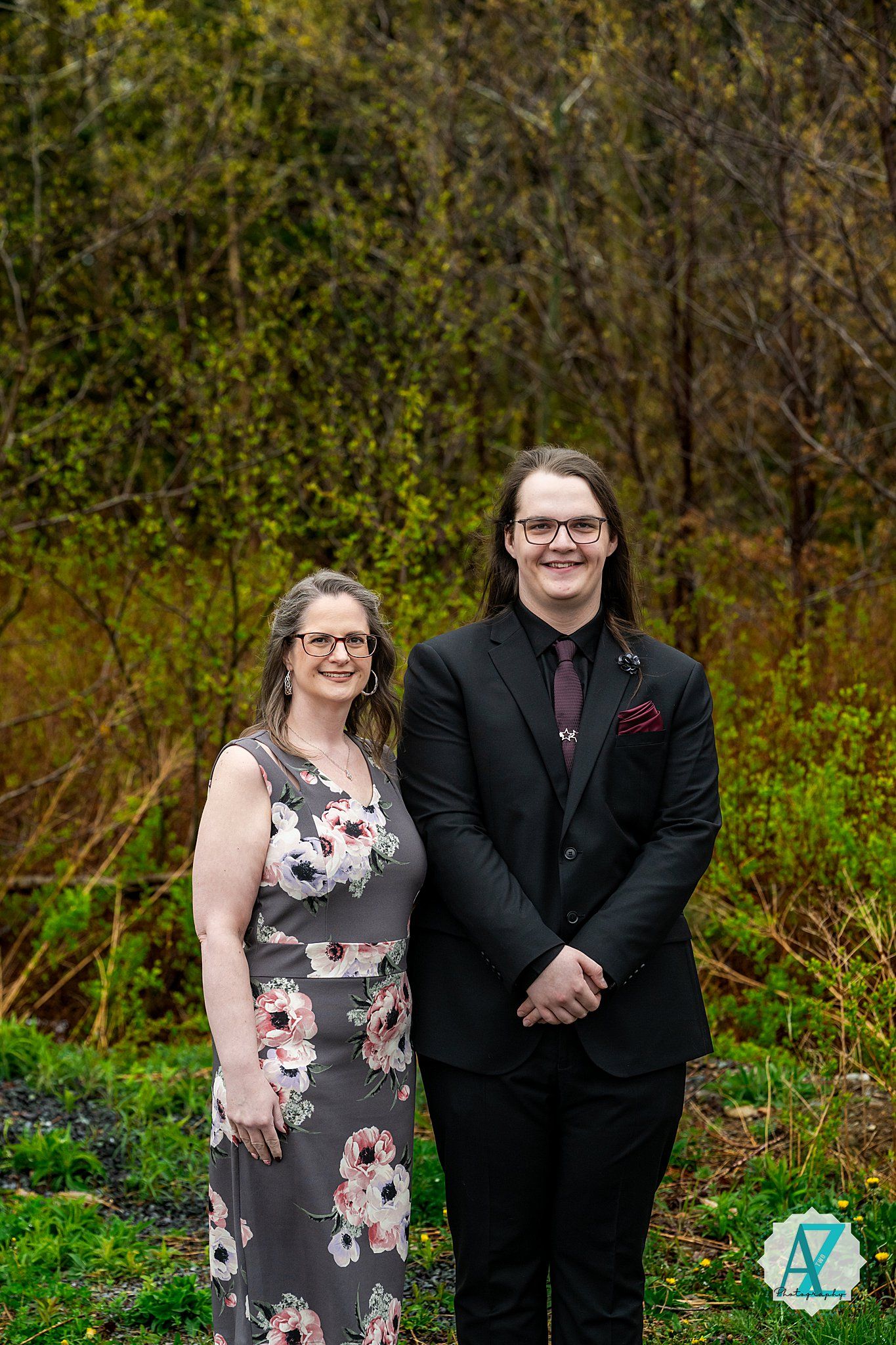 A formal couple stands together outdoors in front of autumn foliage, wearing a dark suit and floral dress.