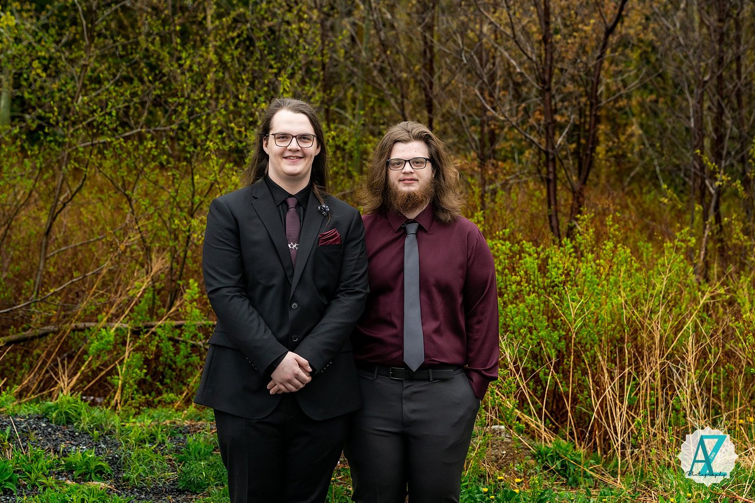 Two people in dark formal attire pose together in front of fall foliage and greenery at an outdoor location.