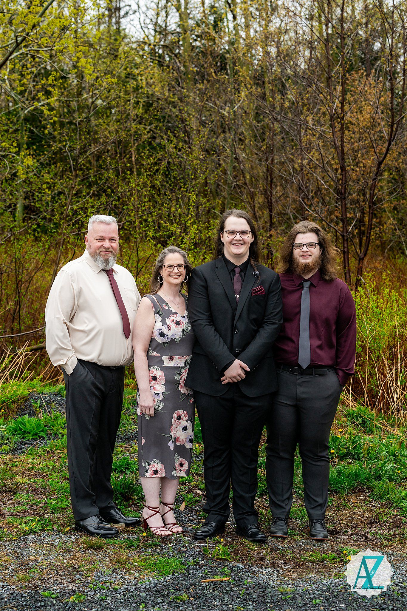 A group of four people wearing formal attire stand together on a gravel path with autumn foliage in the background.