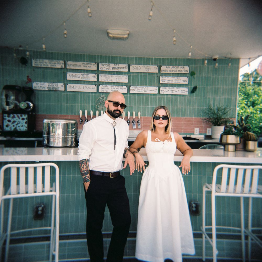 A couple poses at a trendy mint-green food counter with metal barstools, dressed in white and black attire.