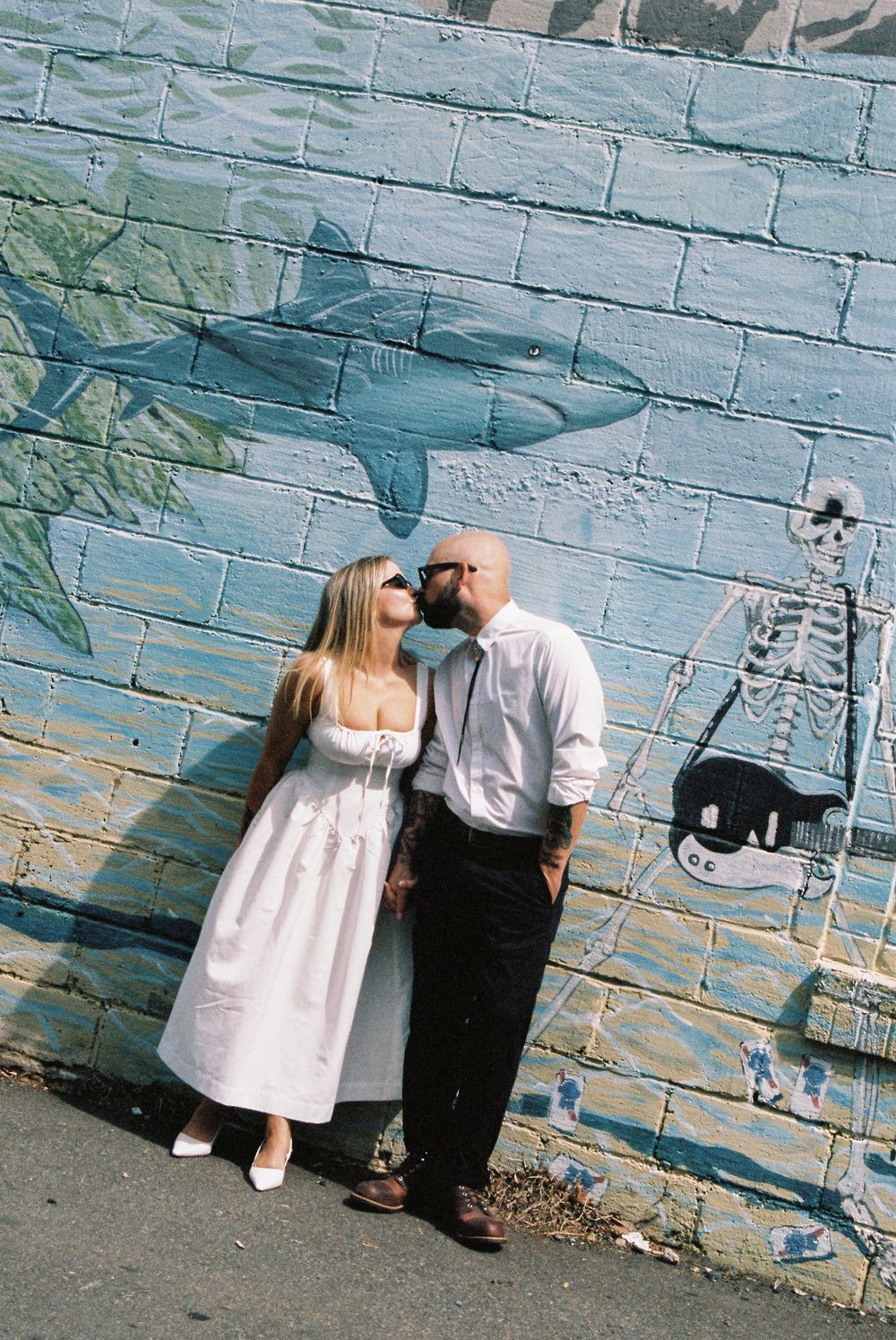 A couple shares a romantic moment against a blue brick wall with painted murals of sharks and guitars.