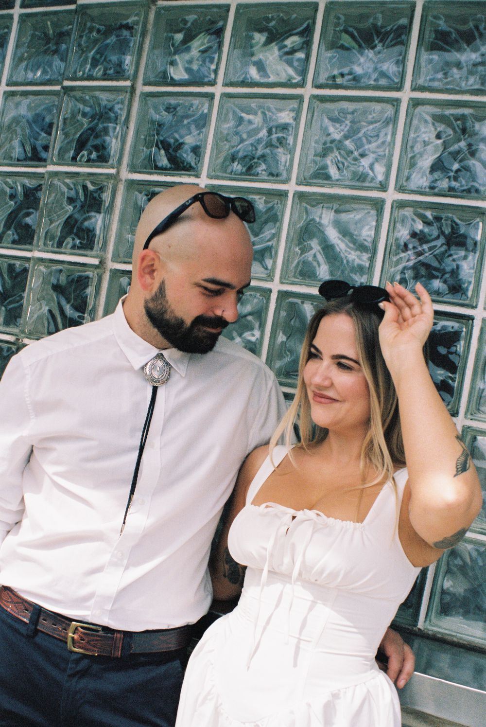 A couple shares a sweet moment against a glass block wall while wearing coordinated white outfits.
