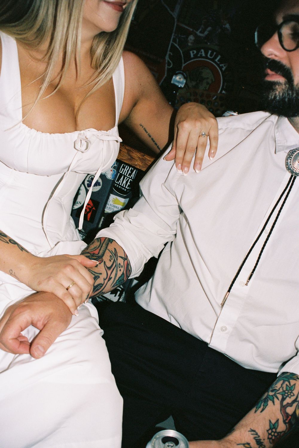 Two people in white outfits sitting close together with tattoos and alcoholic beverages visible in the image.