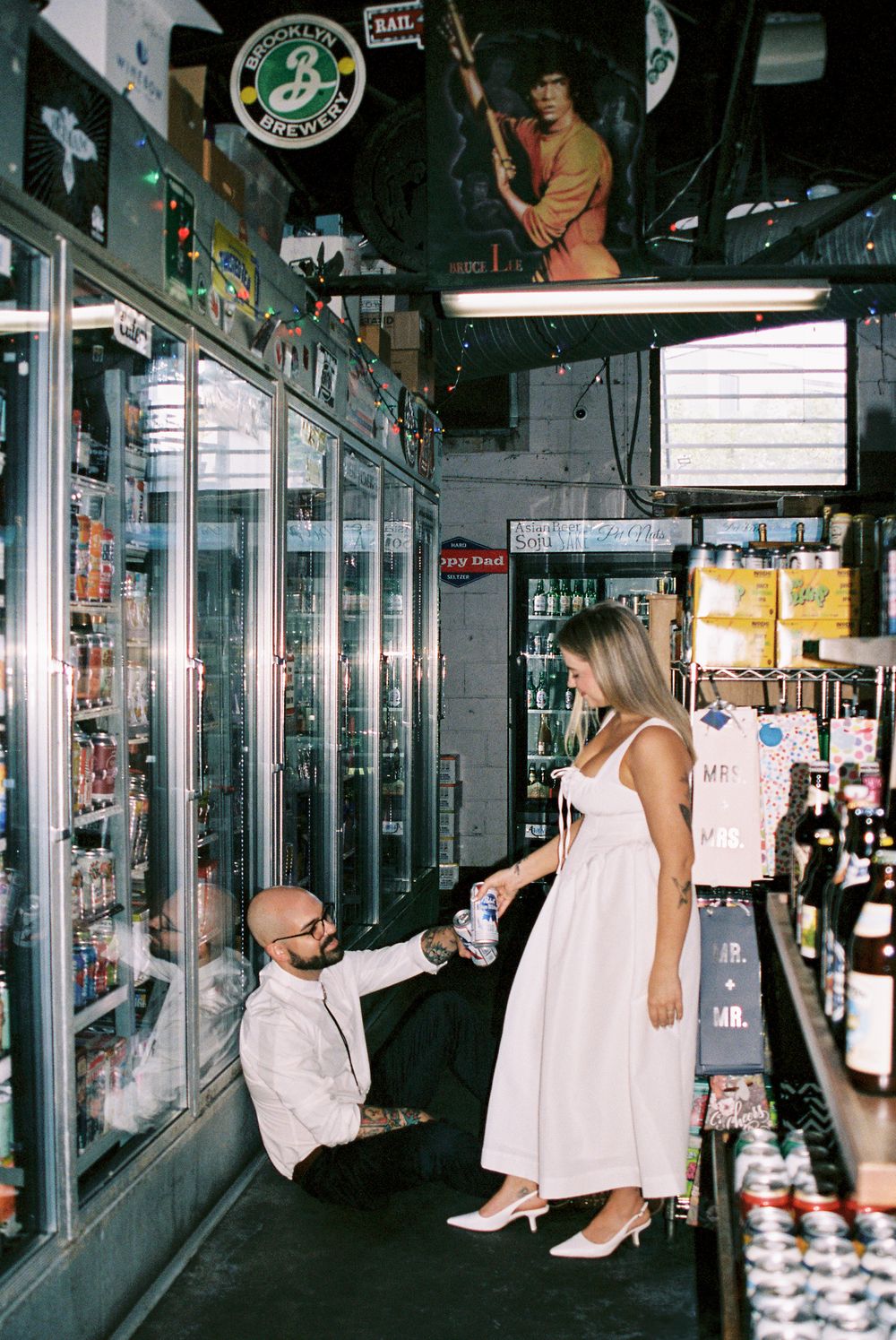 A couple poses together in a convenience store aisle with glass refrigerator doors and a Brooklyn Brewery sign visible.