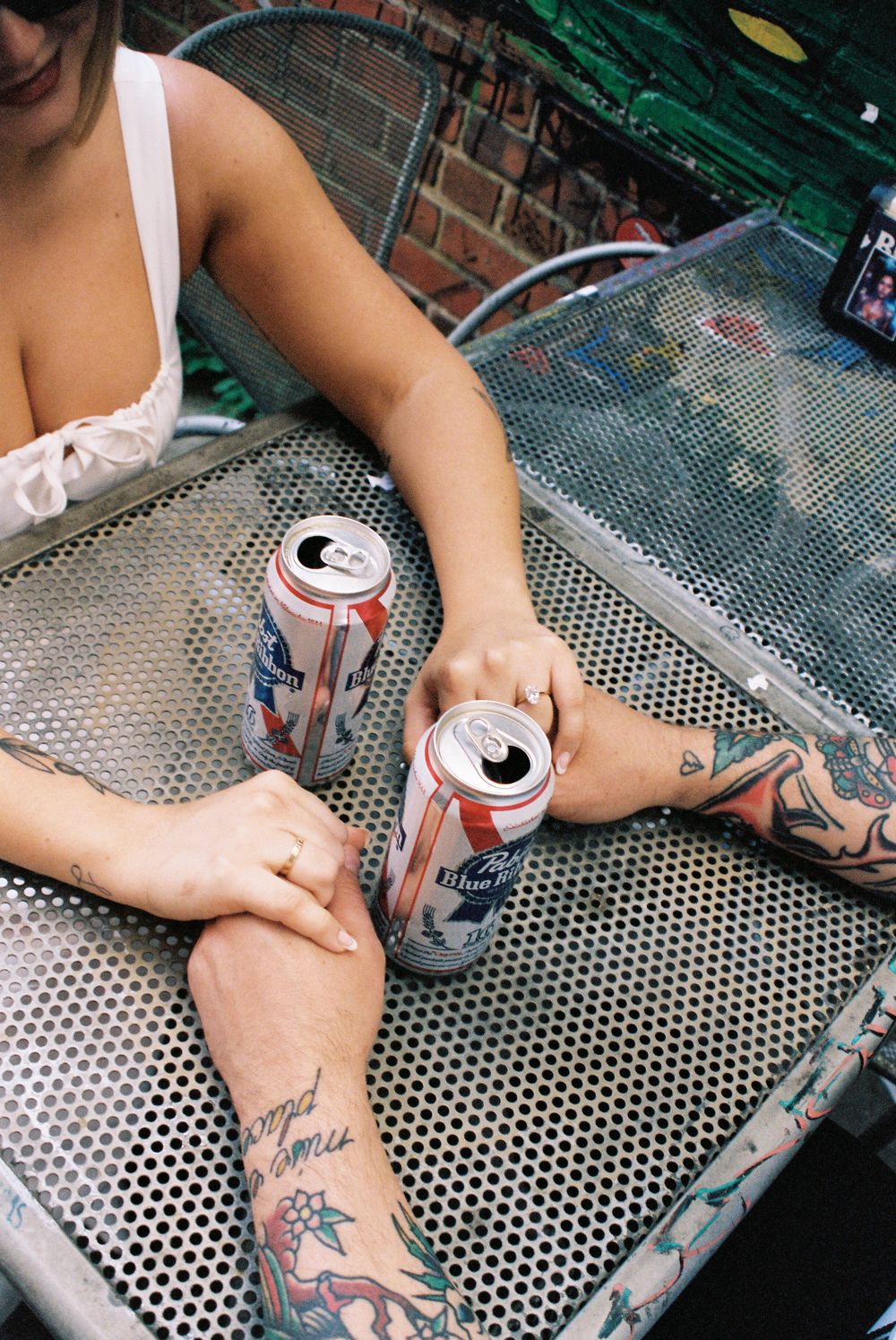 Two cans of beer sit on a metal patio table with visible tattoo art on person's arms.