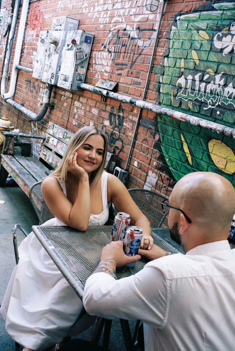 Two people share drinks at an outdoor table against a brick wall with graffiti and street art in an urban setting.