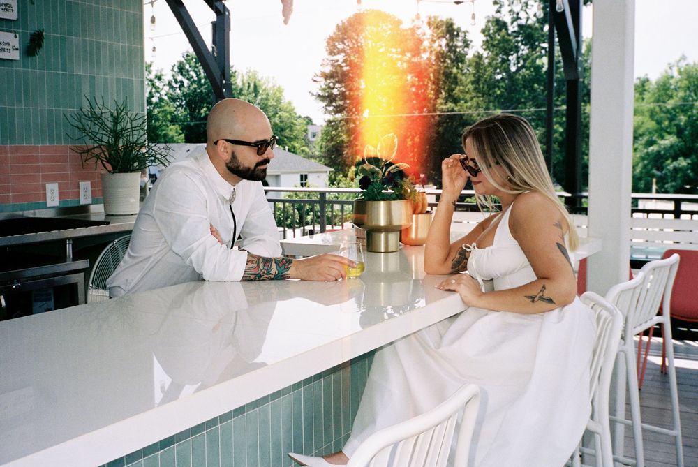 Two people having drinks on an outdoor patio with golden sunlight filtering through trees at sunset.