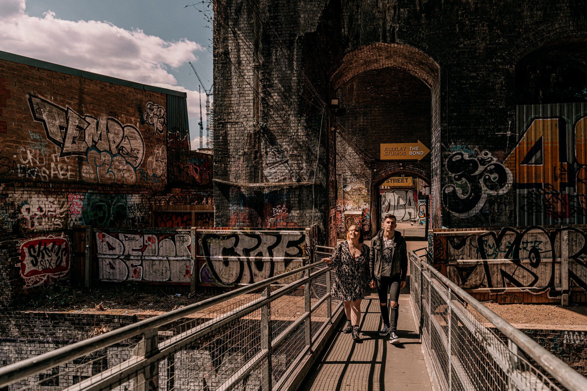 Urban graffiti covered walkway with metal railings under bridge in moody industrial setting at sunset.