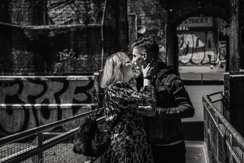 A romantic black and white street scene shows a couple embracing near graffitied walls in an urban setting.