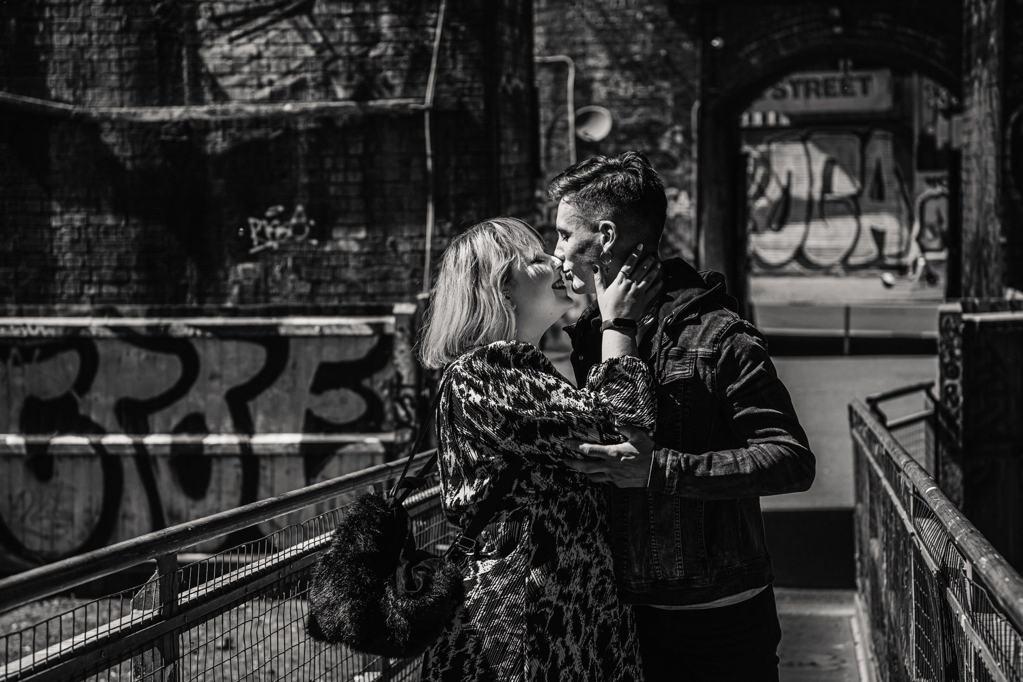 A romantic black and white street scene shows a couple embracing near graffitied walls in an urban setting.