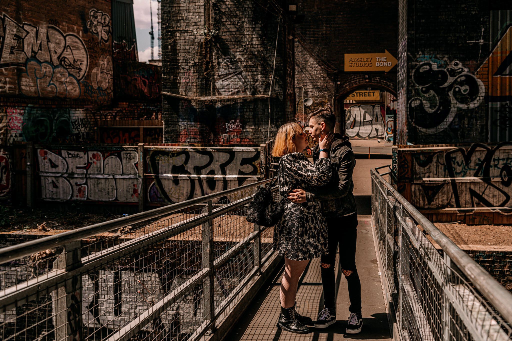 A couple embraces on an industrial walkway covered in graffiti and urban art.