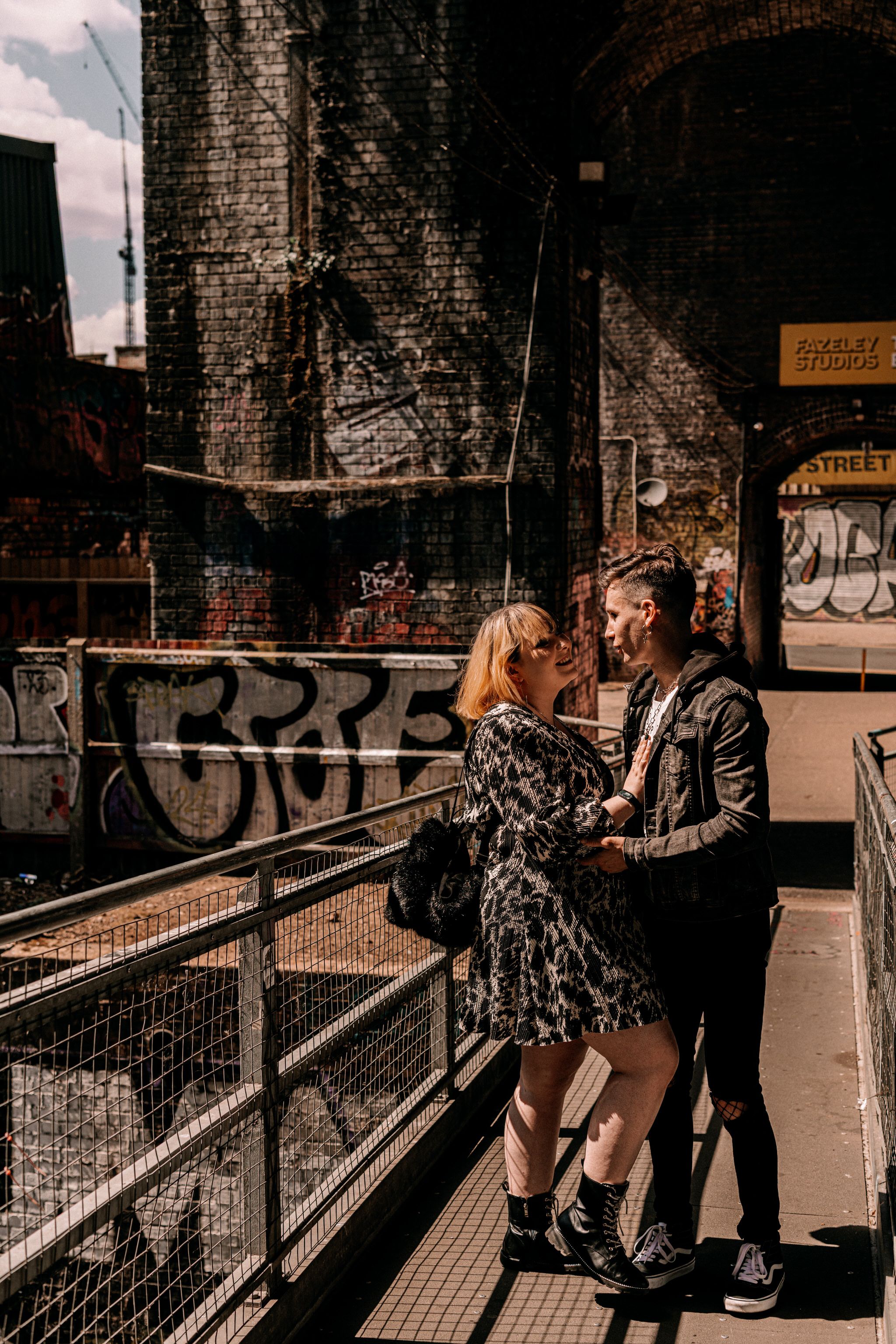 Couple in urban setting standing by brick wall with graffiti while enjoying an intimate moment on industrial walkway.