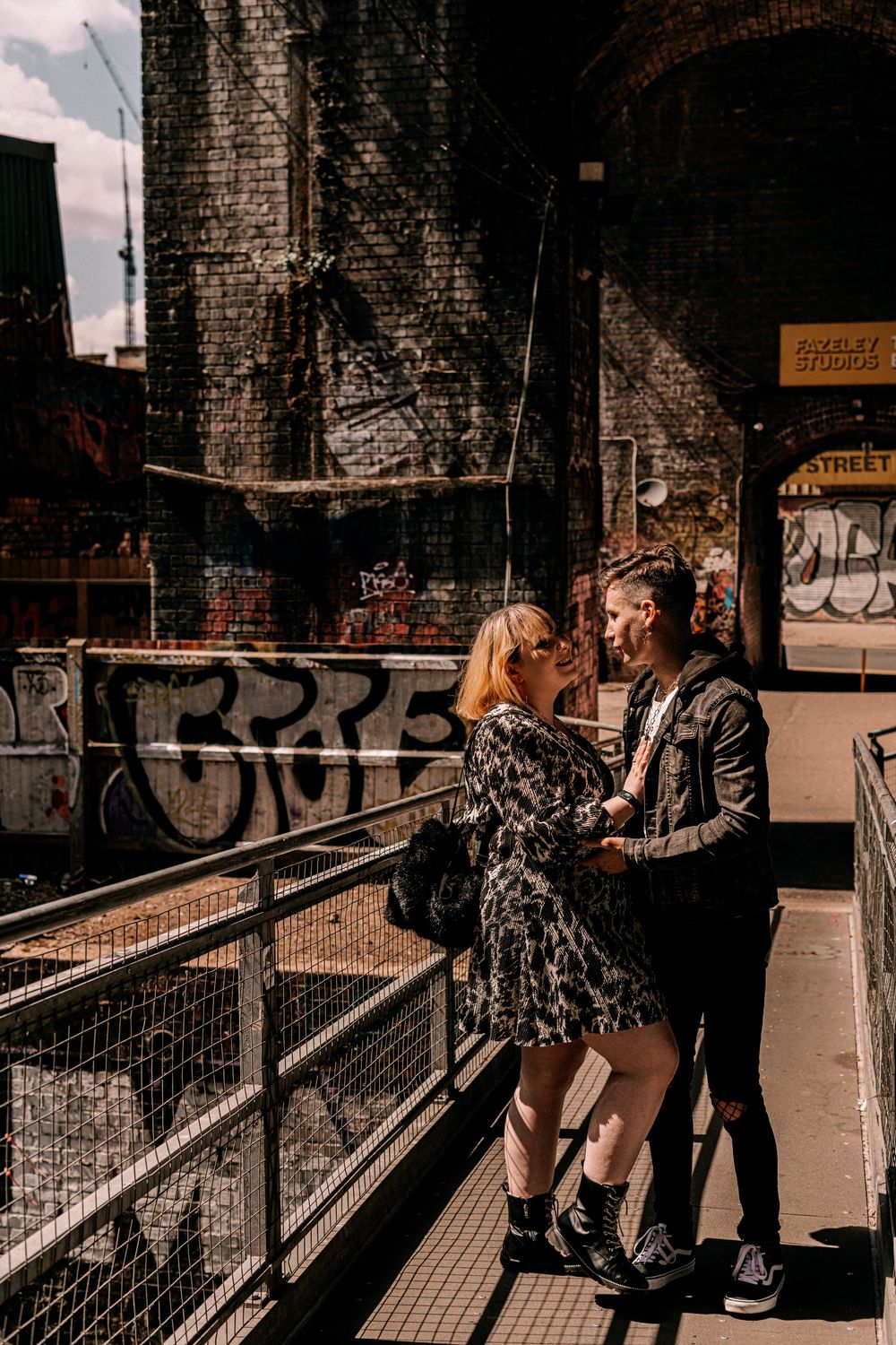 Couple in urban setting standing by brick wall with graffiti while enjoying an intimate moment on industrial walkway.