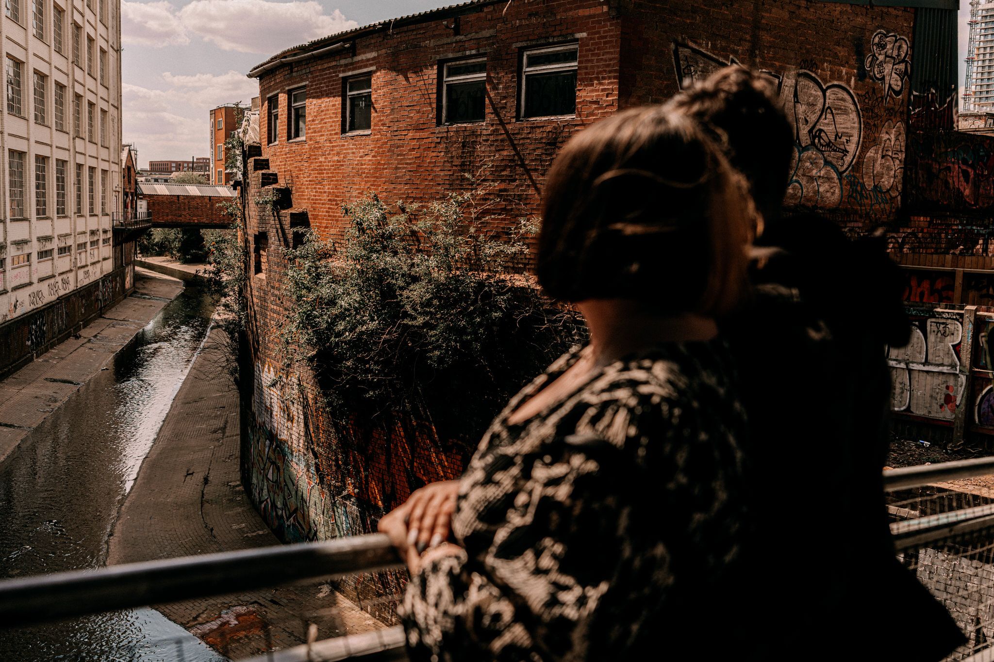 A silhouetted figure overlooks an urban scene with brick buildings and train tracks on an overcast day.