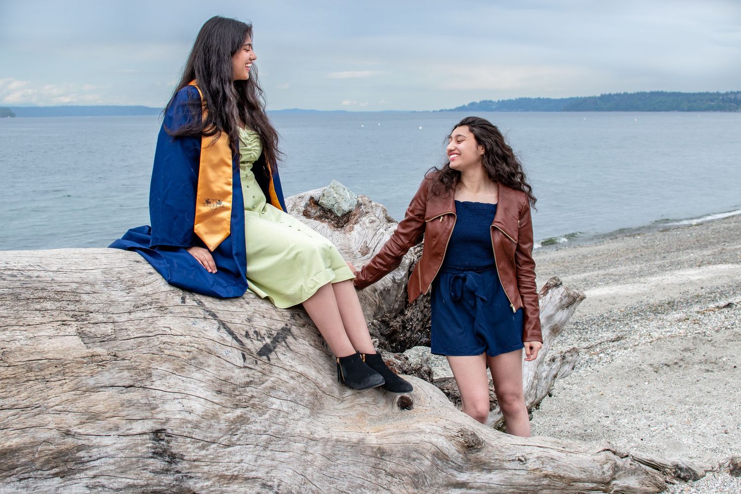 Sisters sitting on driftwood at Redondo Beach, sharing laughter during a lighthearted spring graduation photo session.