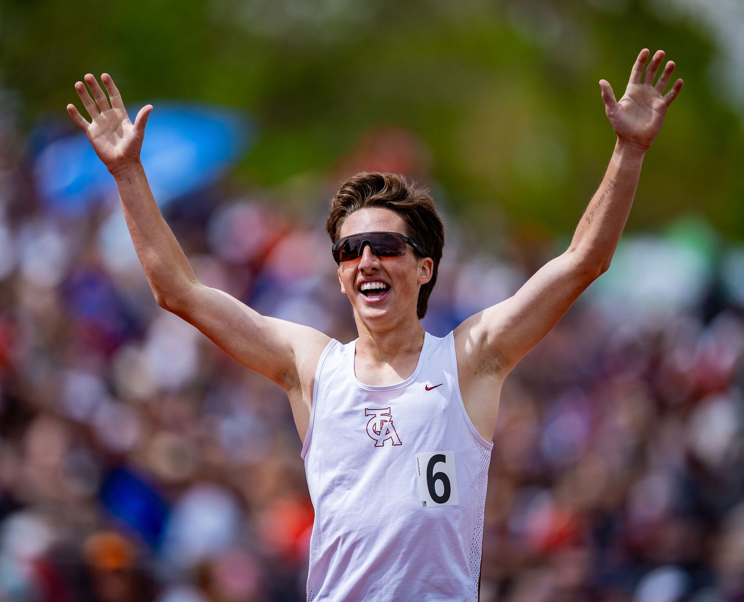 Athlete wearing sunglasses and white shirt with number 6 raises arms in celebration during outdoor competition.