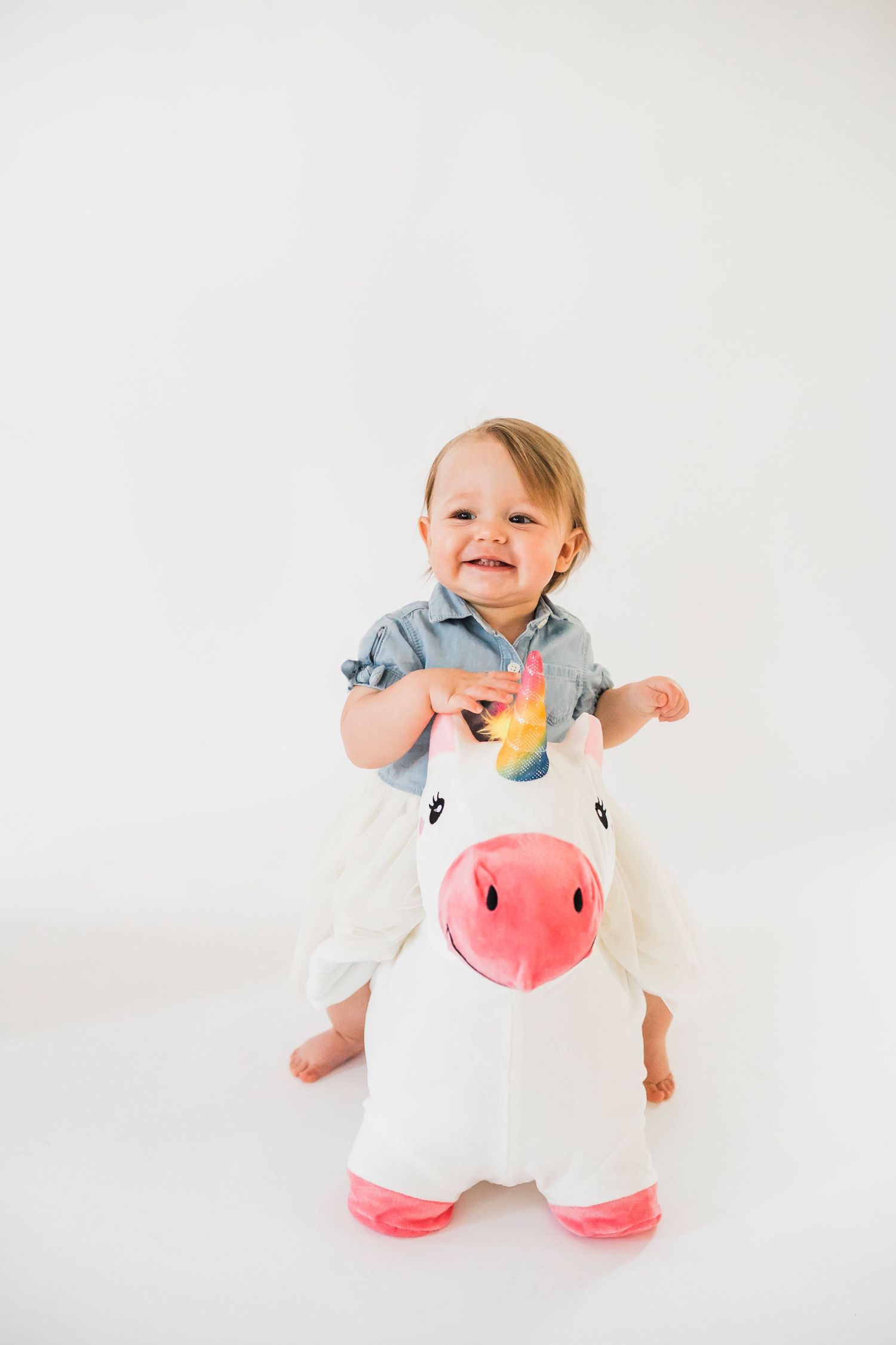 Young child in denim shirt and white pants sits atop a plush white unicorn toy with pink accents against white backdrop.