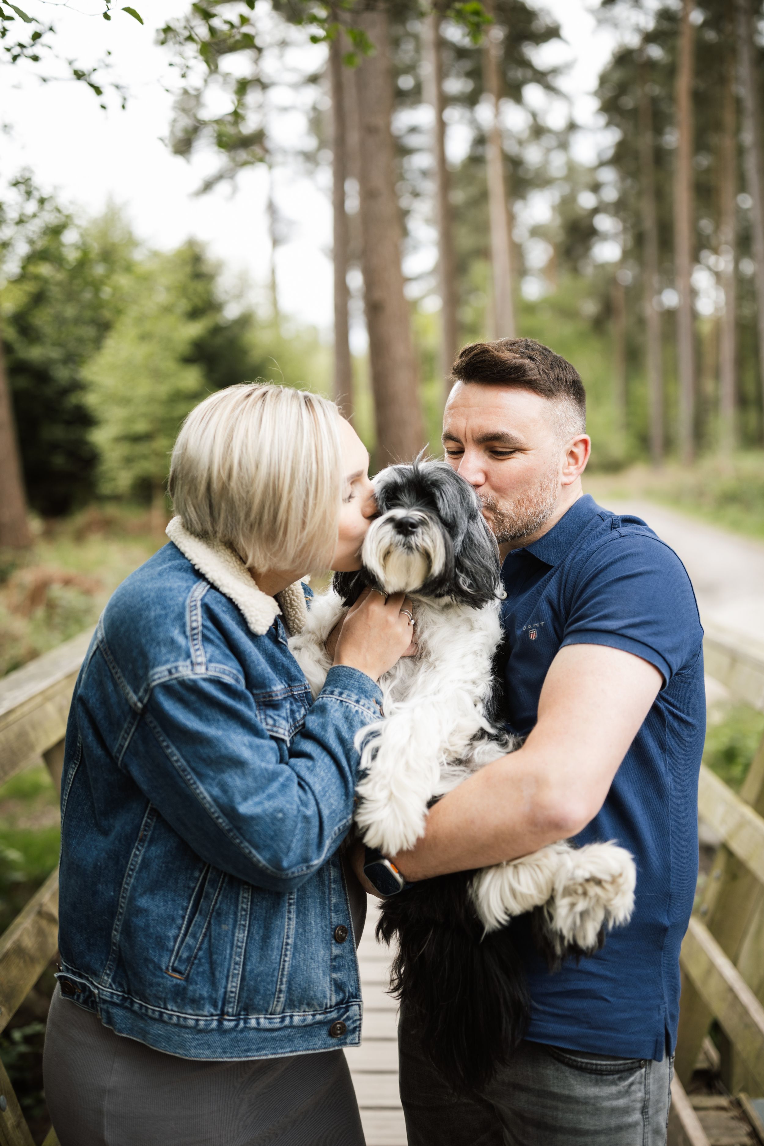 A Shih Tzu puppy receives affectionate kisses from its owners on a wooden walkway in a forest setting.