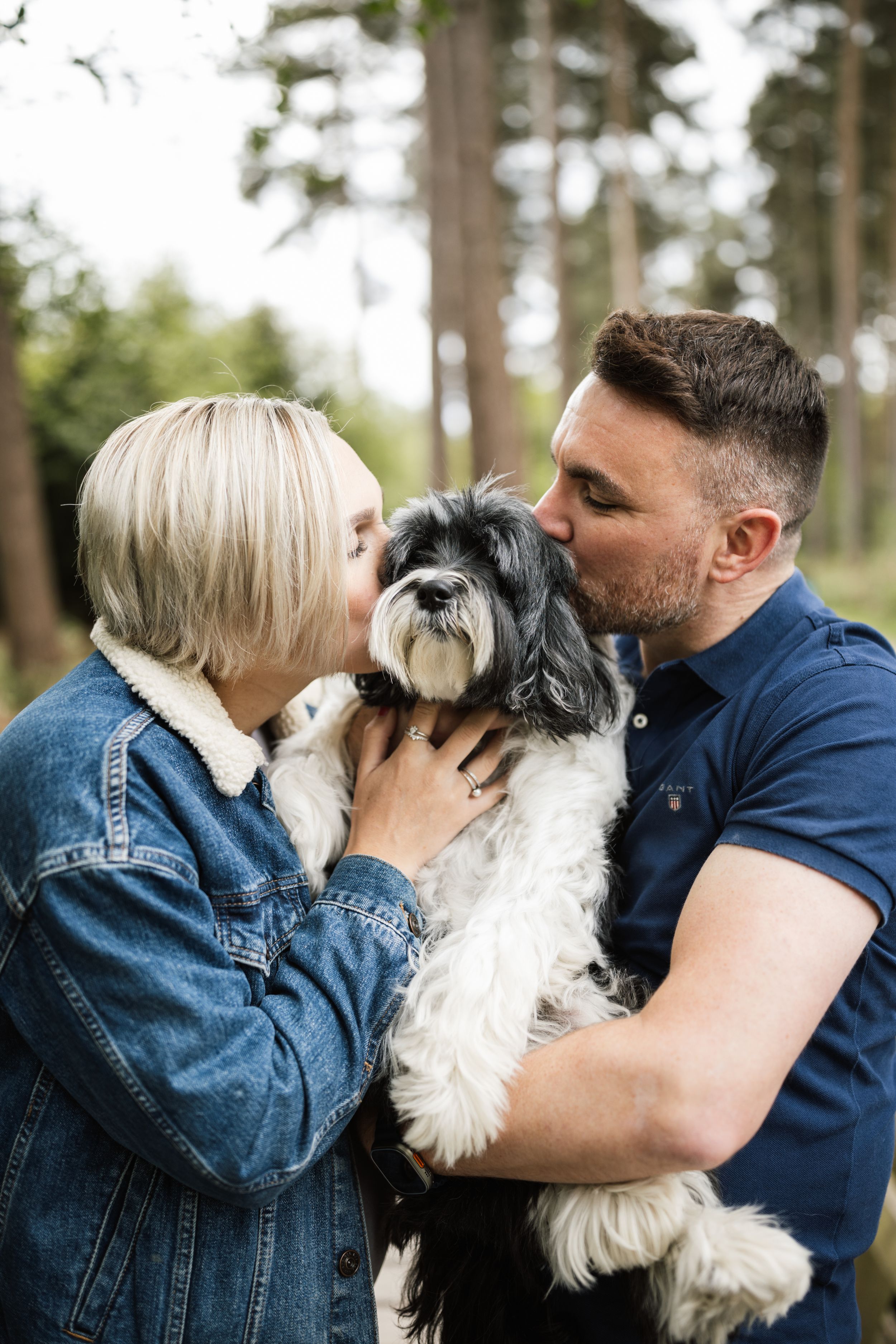 A black and white Shih Tzu dog being lovingly embraced by two people in a forest setting.