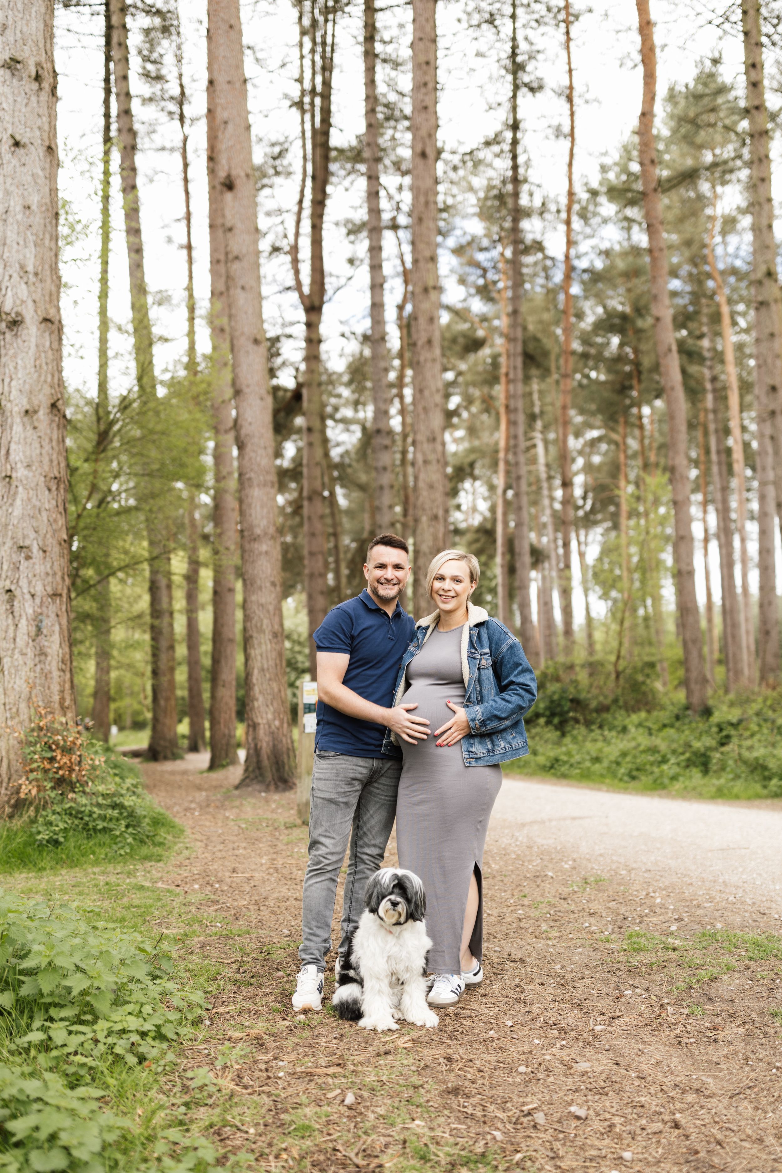 A couple with their black and white dog pose together on a forest trail during a maternity photoshoot.