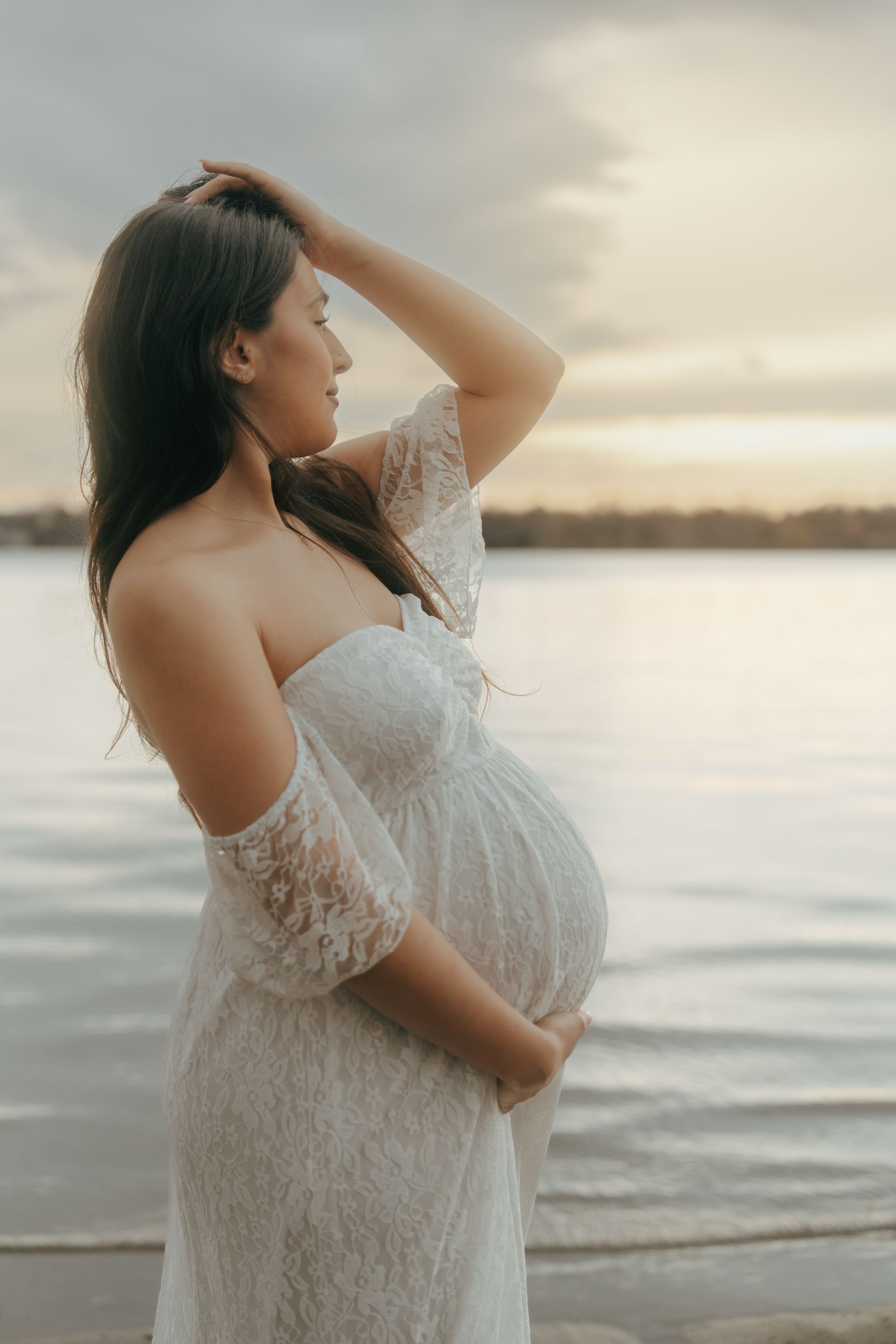 A pregnant woman in a white lace dress poses on a beach at sunset, cradling her baby bump.