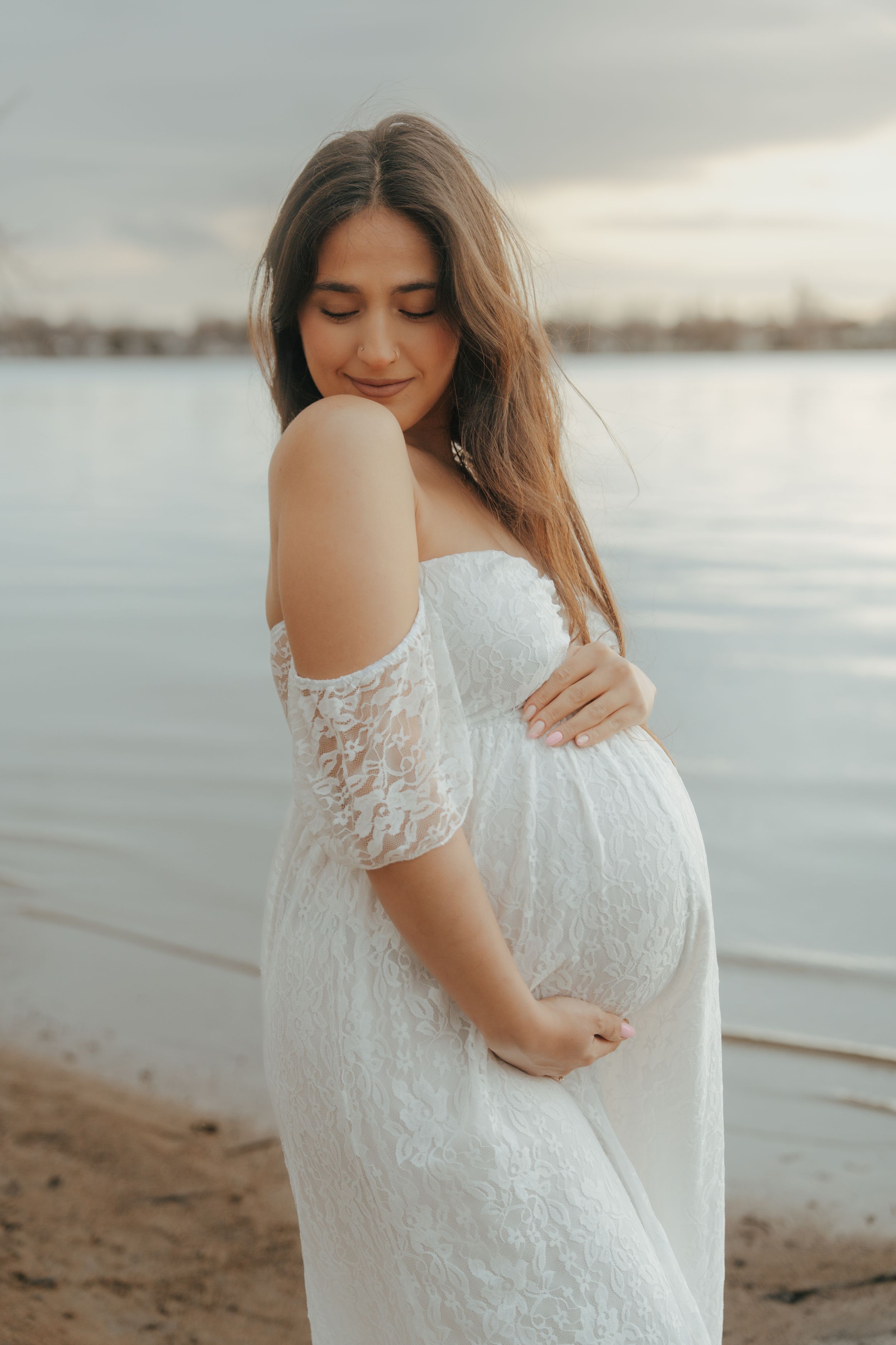 A person in a white lace maternity dress poses tenderly at the beach during sunset.
