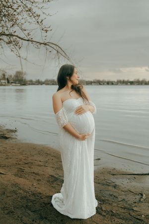 An expectant woman in a white flowing dress poses thoughtfully by a peaceful lakeside at sunset.