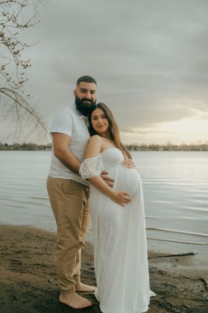 Expectant couple embracing by a lake at sunset during maternity photoshoot.