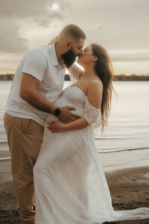 A romantic sunset beach photo of an expecting couple in white sharing an intimate moment by the water.