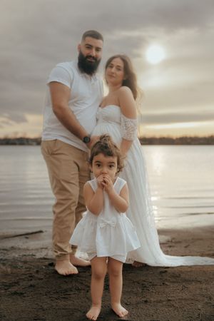 A family poses for a maternity photo on the beach at sunset wearing coordinated white outfits.