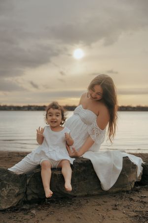 Two people in white dresses sit on rocks by a lake during sunset with cloudy skies creating a dreamy atmosphere.