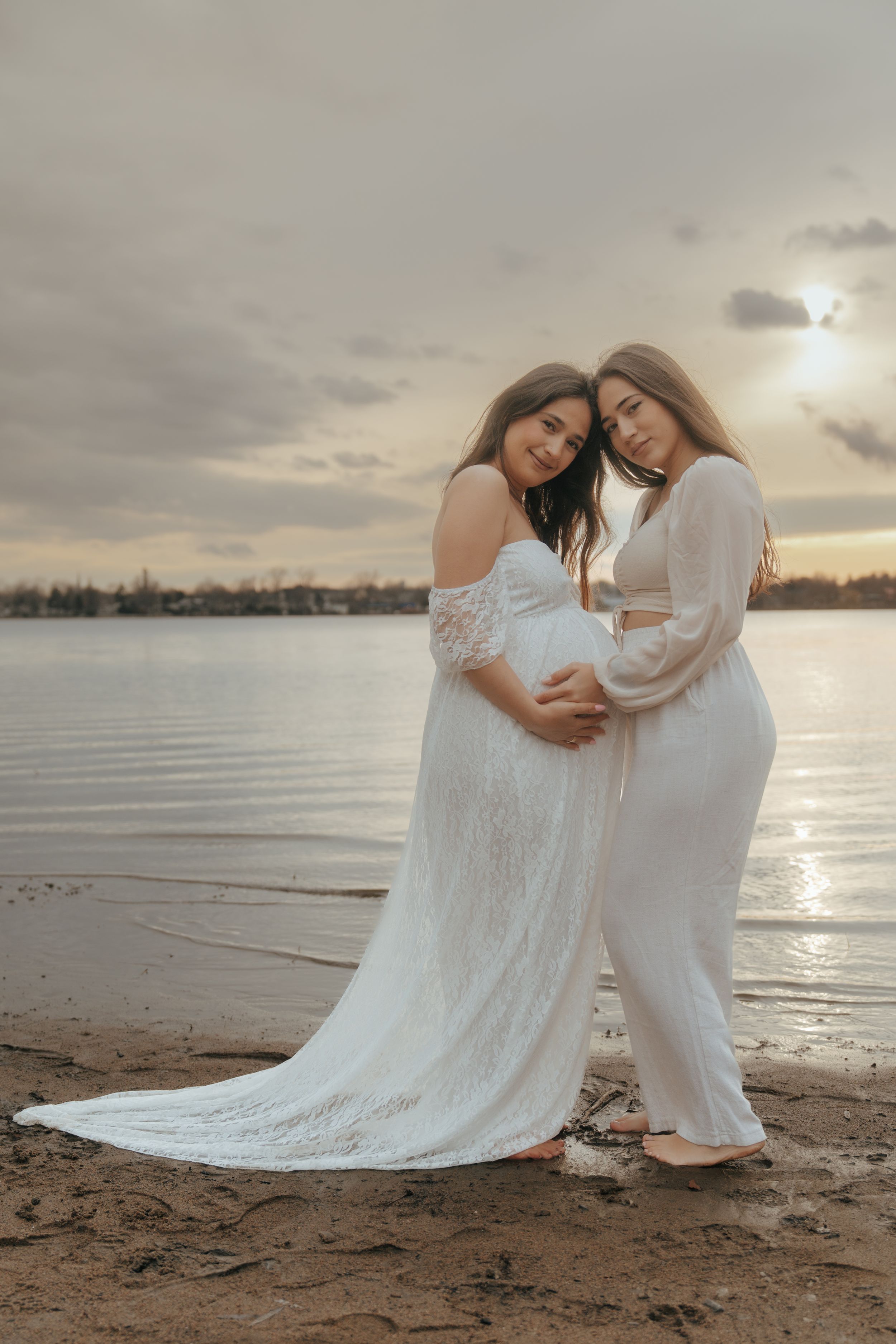 Two women in white dresses embrace on a beach at sunset creating an intimate maternity moment.