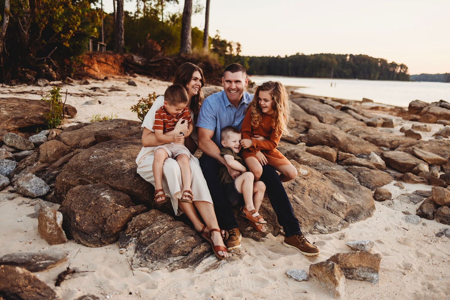 A group relaxes together on rocky coastal shoreline at sunset with pine trees and water in the background.