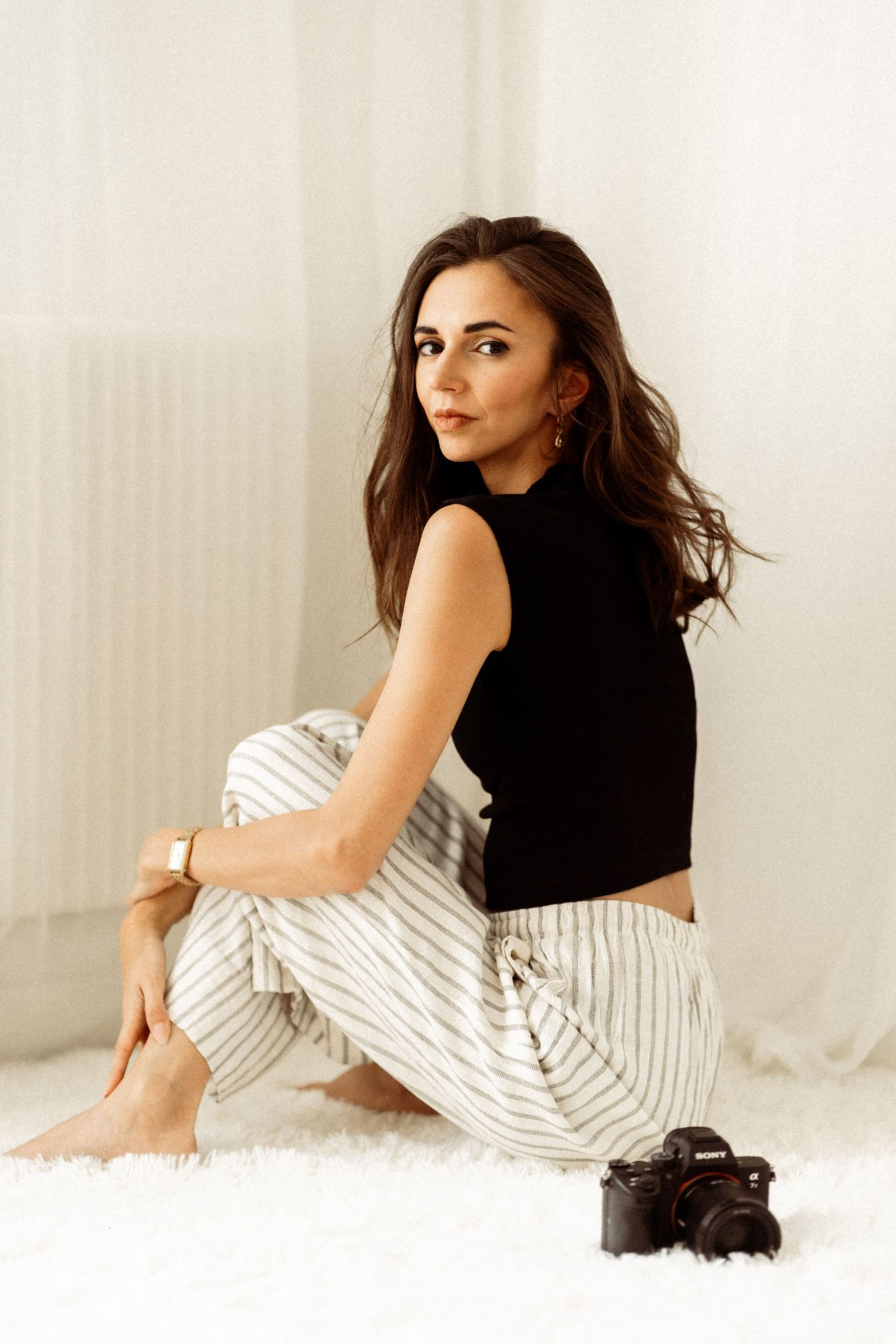 Model in black crop top and striped pants poses on floor with camera nearby in bright interior setting.