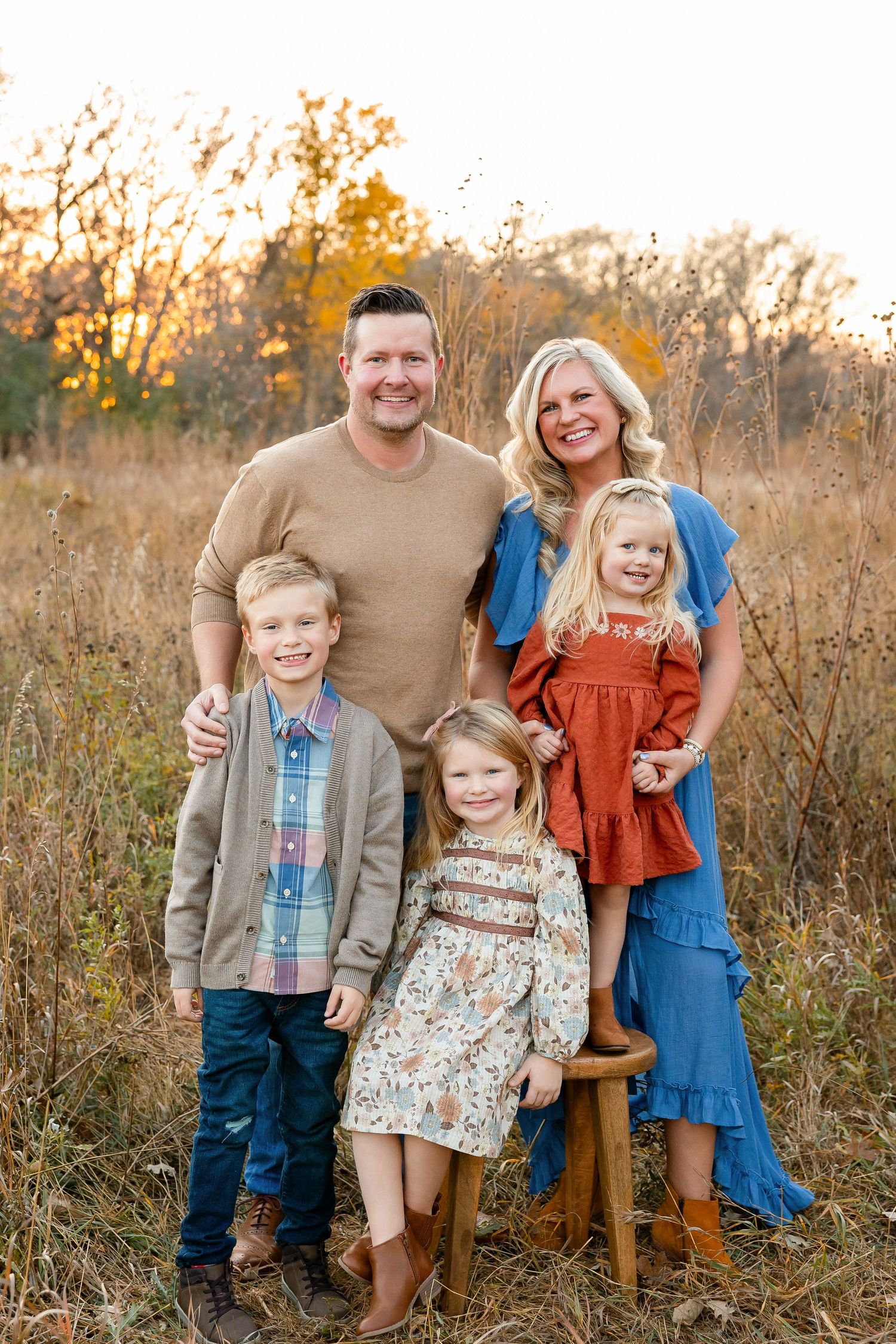 A family sits together on a blanket in a golden autumn field with evergreen trees and red foliage in the background.